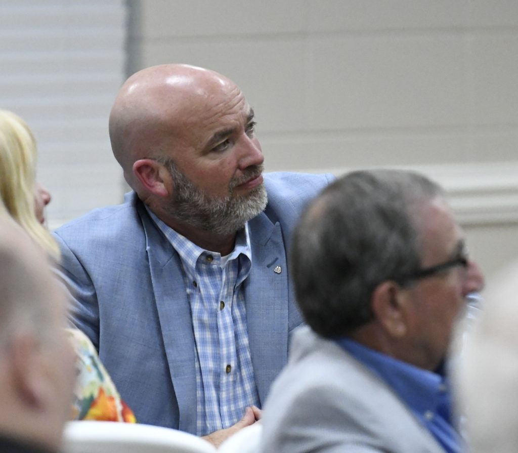 Bald man with a beard wearing a blue blazer and plaid shirt listens attentively at an indoor event, with other people seated around him.