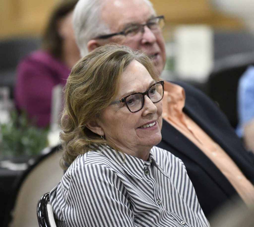 Older woman with glasses smiling, seated at an indoor event, wearing a striped shirt. Out-of-focus man and other attendees sit behind her.