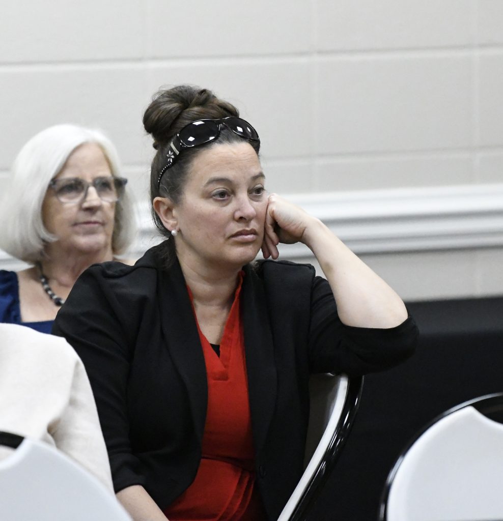 Two women sitting indoors, one in the foreground with sunglasses on her head looking pensive, and another in the background with white hair and glasses.