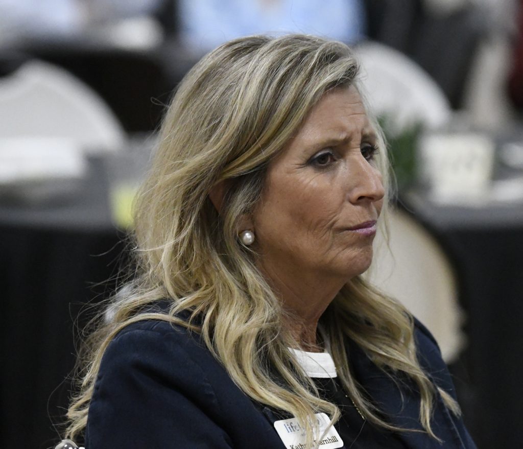 A woman with long blonde hair sits indoors, looking thoughtful. She wears a dark jacket, pearl earrings, and a name tag. The background is blurred.
