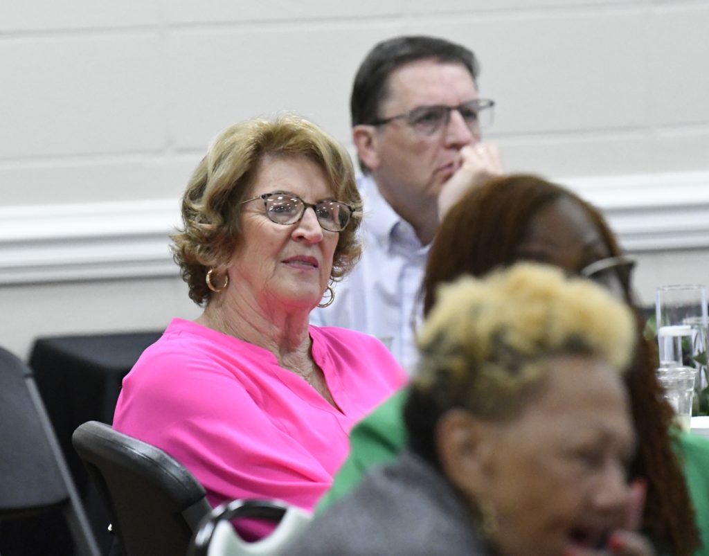 A woman in a bright pink blouse sits attentively at an event, with other people seated around her in a conference or meeting room.