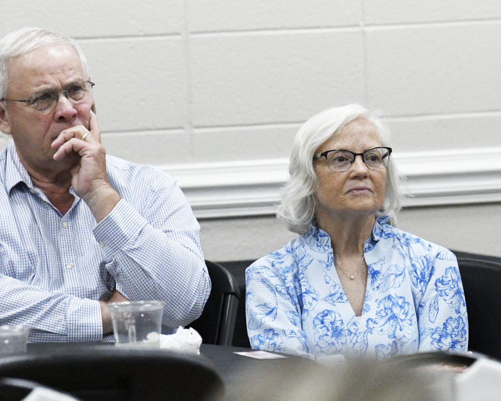older man and woman with glasses sit at a table, appearing thoughtful and attentive, in a brightly lit room with light-colored walls.
