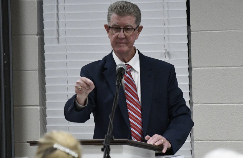 A man in a suit and striped tie speaks at a podium with a microphone, gesturing with his hand, in an indoor setting with white blinds behind him.