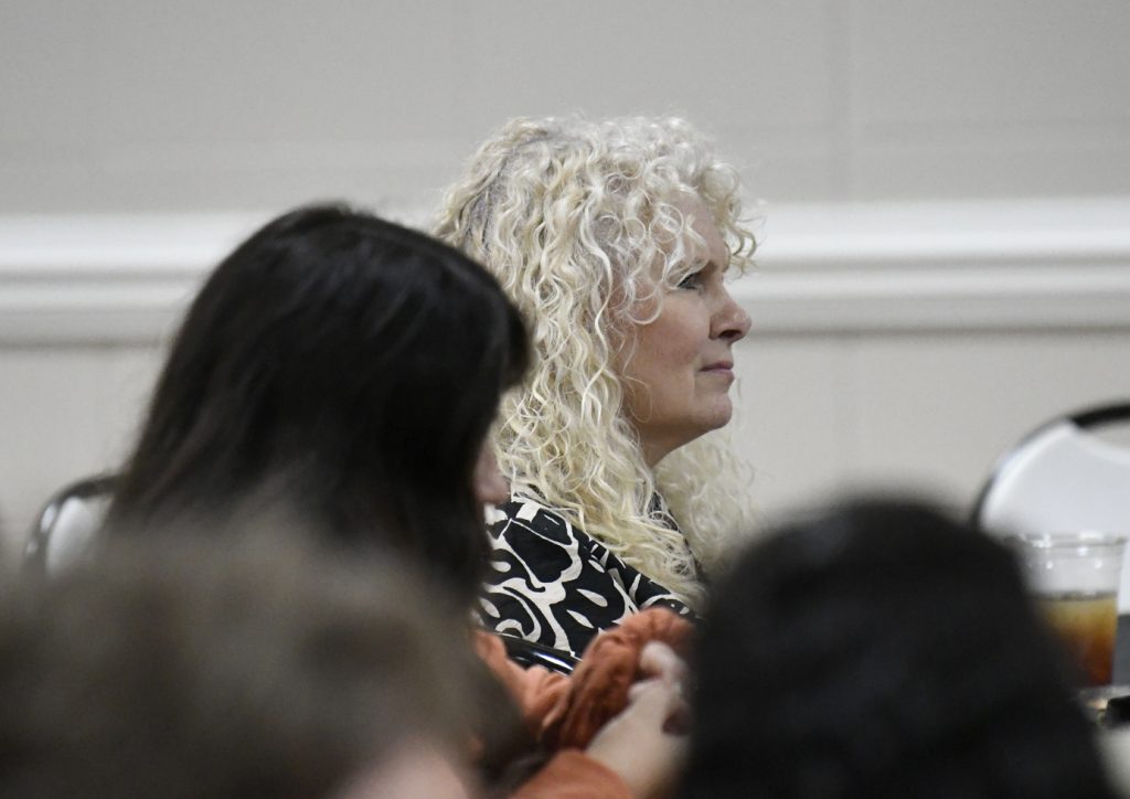 A woman with curly blonde hair attentively listening at an indoor event, with other people partially visible in the foreground.