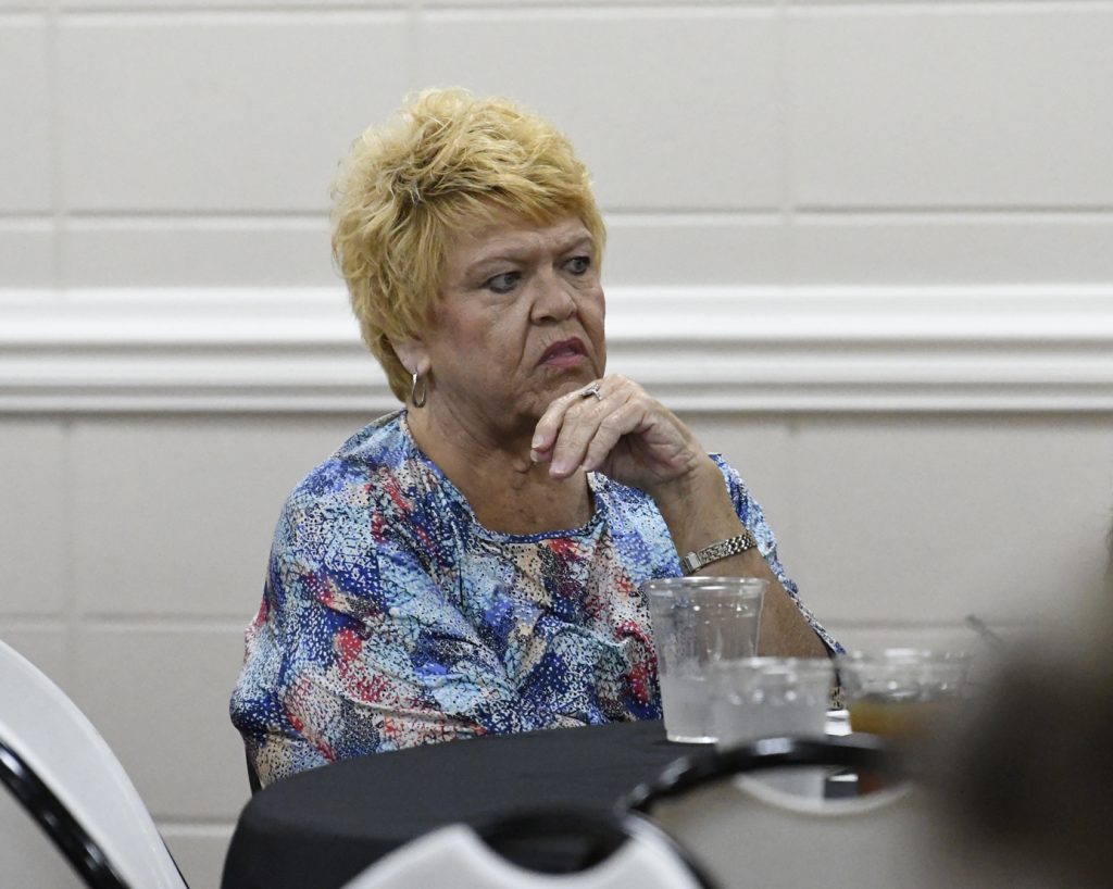 Older woman with short blonde hair sits at a table, looking thoughtful, with hand near chin and empty cups in front of her.