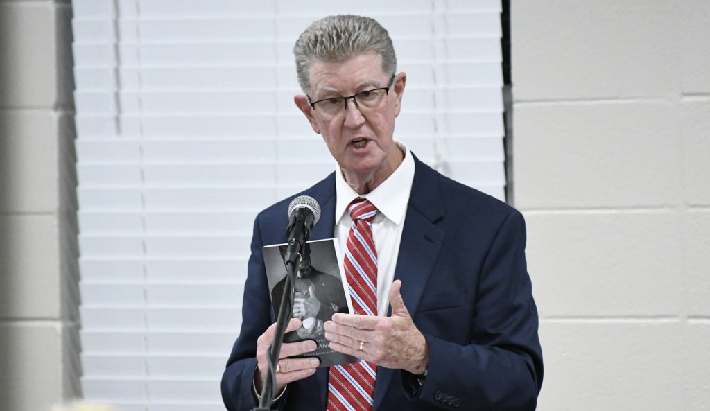 Man in suit and striped tie speaks into a microphone, holding up a book in front of window blinds.