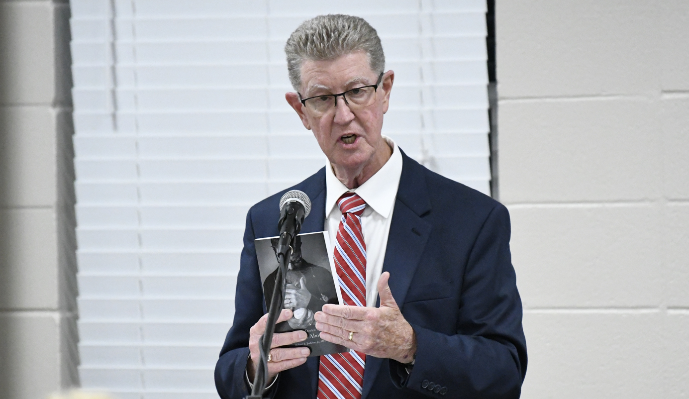 Man in suit and striped tie speaks into a microphone, holding up a book in front of window blinds.