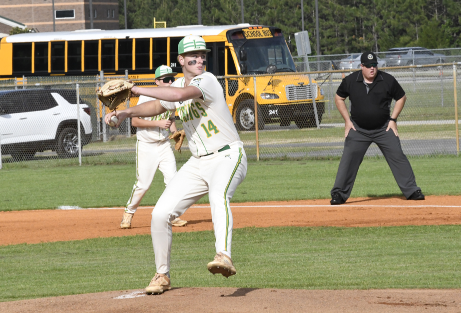 Baseball pitcher in white uniform throwing a pitch, with teammates and umpire on the field. School bus and cars are visible in the background.