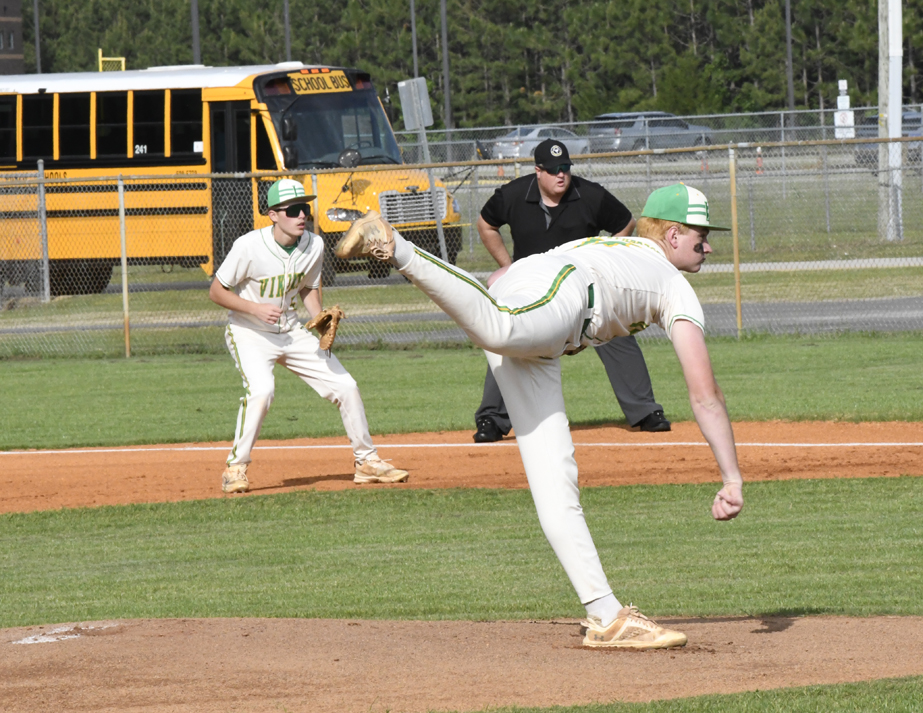 A baseball pitcher throws the ball during a game, with an infielder ready and an umpire watching. A yellow school bus is visible in the background.