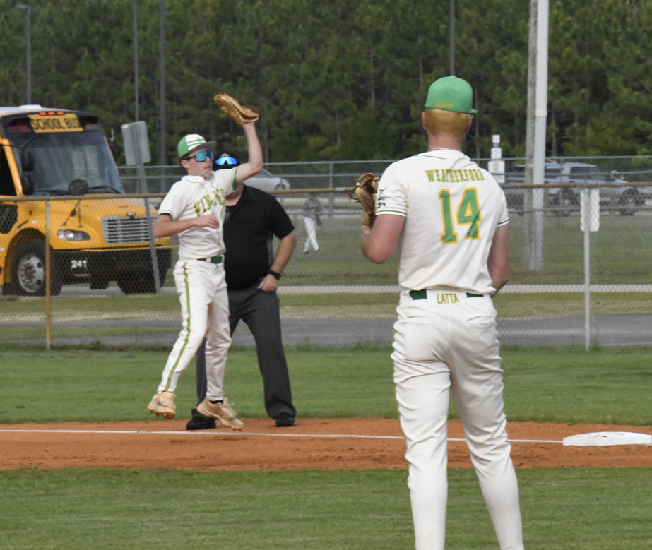 Two baseball players in white uniforms and green caps are on a field, with an umpire, school bus, and fence in the background.