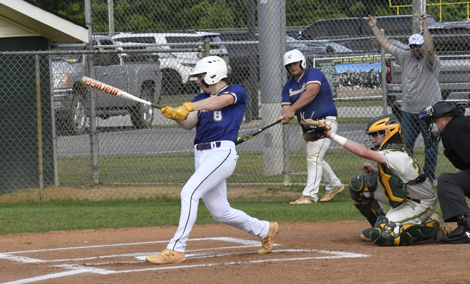 Baseball player in blue uniform swinging bat, catcher and umpire in position, runner on deck, and spectator raising arms behind fence in background.