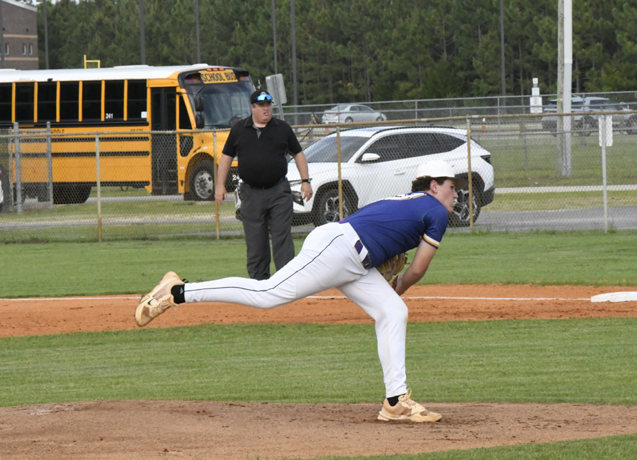 A baseball player in a blue jersey pitches on a field, with an umpire, school bus, and cars in the background.