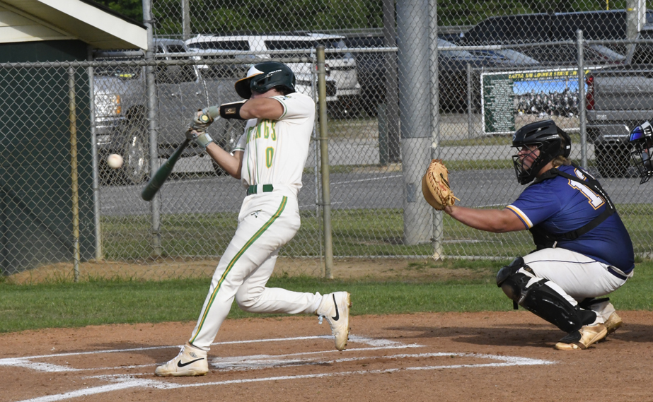 Baseball player in white uniform swinging at a pitch, catcher in blue gear ready to catch the ball, chain-link fence and cars in the background.
