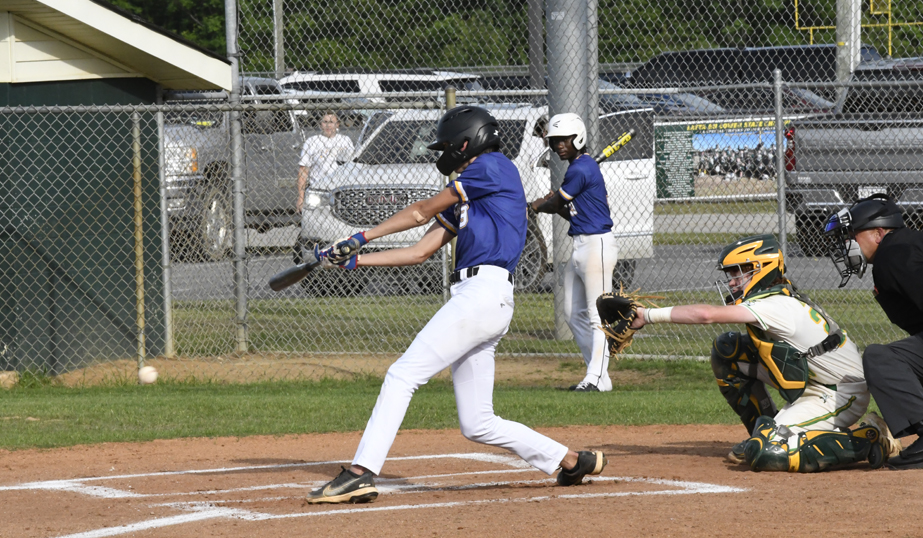 A baseball player swings at a pitch during a game, with a catcher, umpire, and another player in the background near a chain-link fence.
