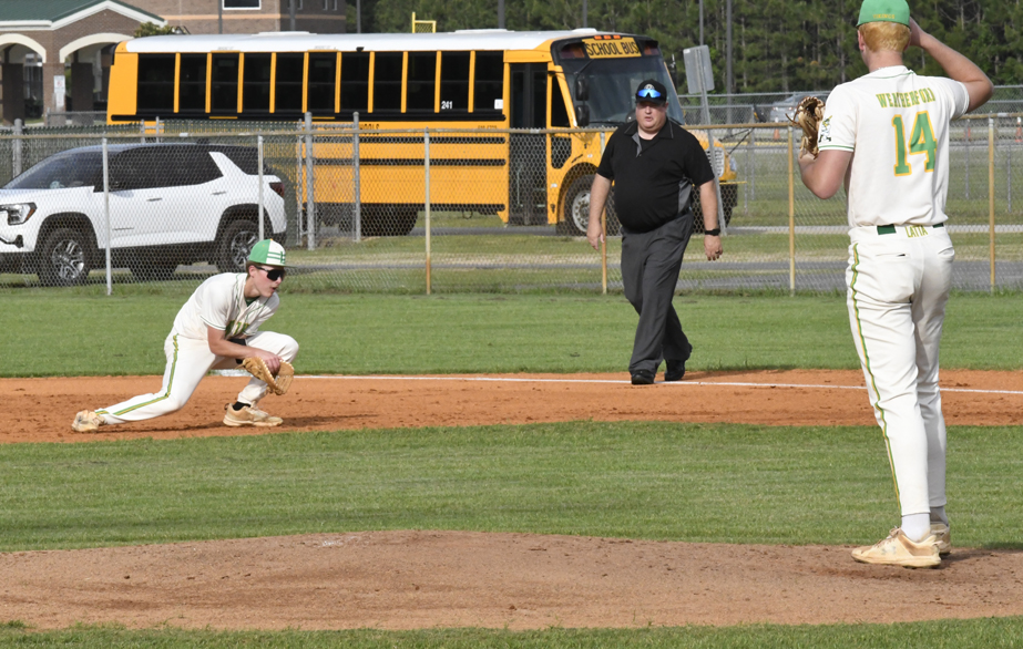 A baseball player prepares to field a ground ball as another player and an umpire watch. School buses and a car are visible in the background.