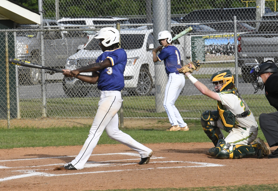 A baseball player in a blue jersey swings at a pitch while the catcher, umpire, and on-deck batter watch, near a chain-link fence with vehicles in the background.