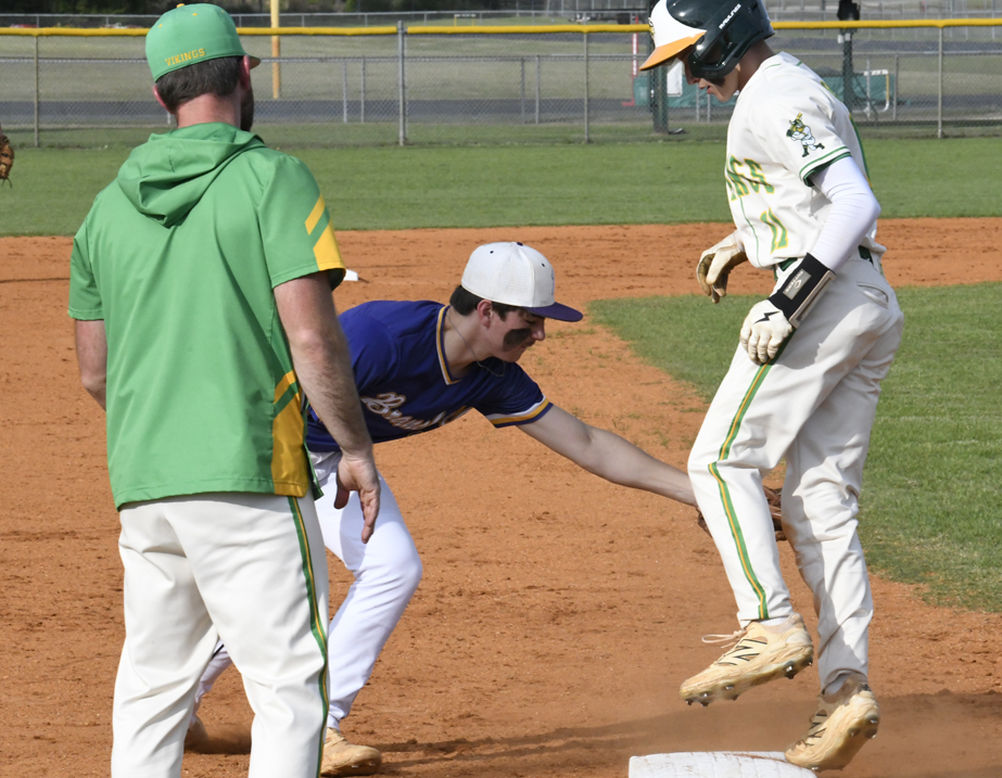 A baseball player in white leaps onto a base as a player in blue attempts to tag him, with a coach in green watching nearby.