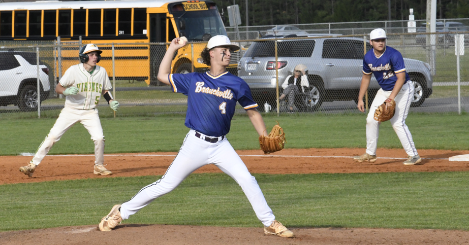 A high school baseball pitcher in blue winds up to throw a pitch while teammates and an opponent stand ready on the field.