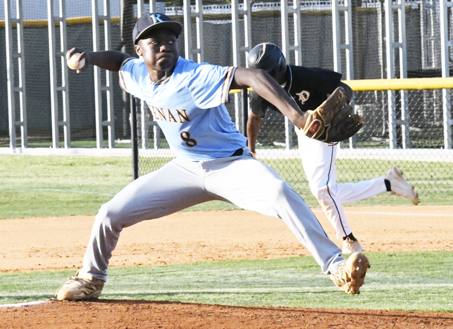 A baseball pitcher in a blue jersey winds up to throw, while a runner in black and white prepares to sprint behind him on the field.
