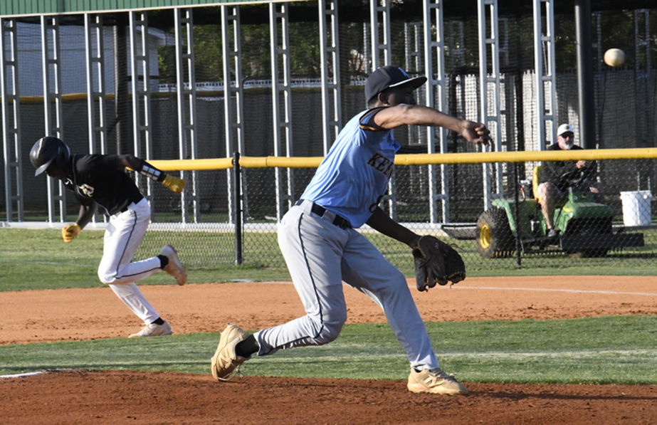 A baseball player in a blue uniform throws the ball as a runner in black sprints toward a base during a game.