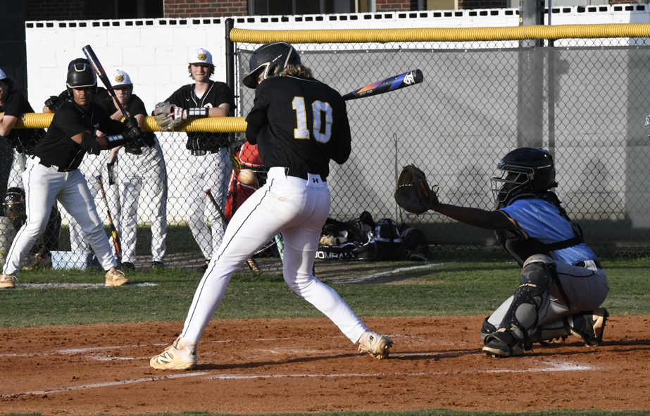 Baseball player in black jersey swinging at pitch, with catcher in gear and teammates watching from behind fence on sunny day.
