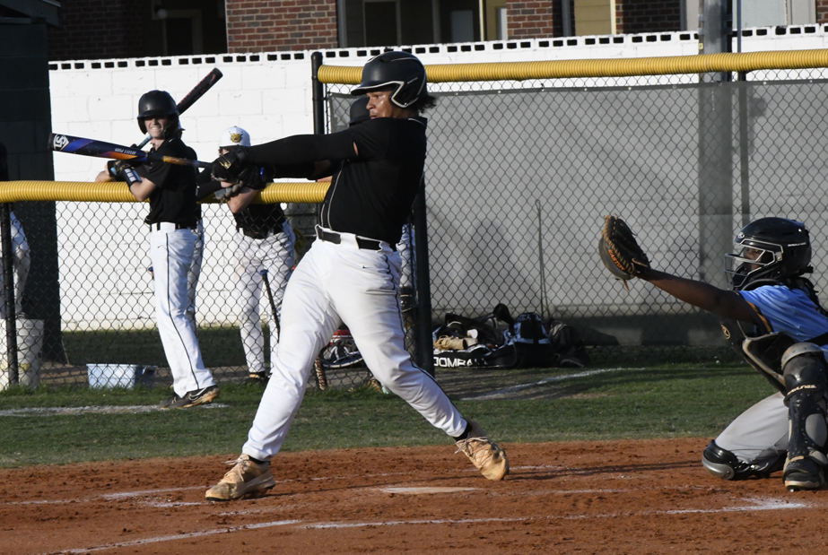 A baseball batter in black uniform swings at a pitch, with a catcher in blue behind him and teammates watching from the dugout area.