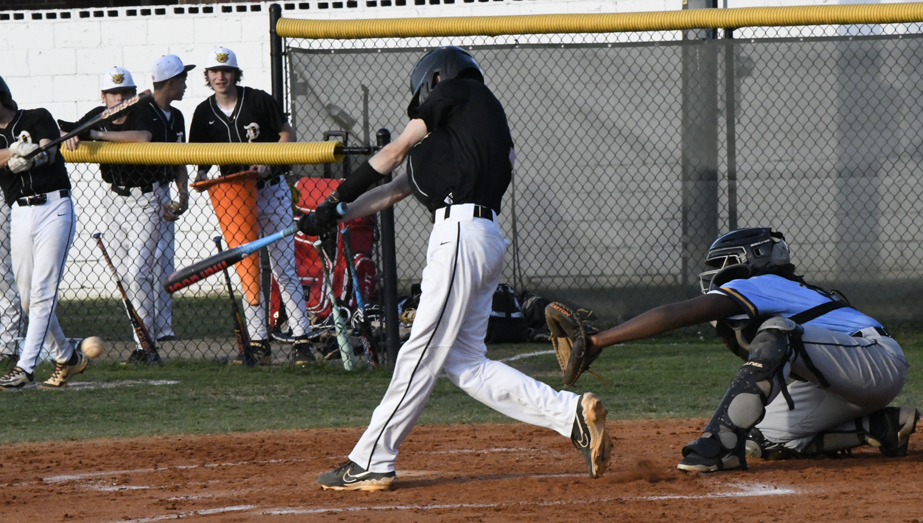 A baseball player in black swings at a pitch while a catcher in blue crouches and teammates watch from behind the fence.