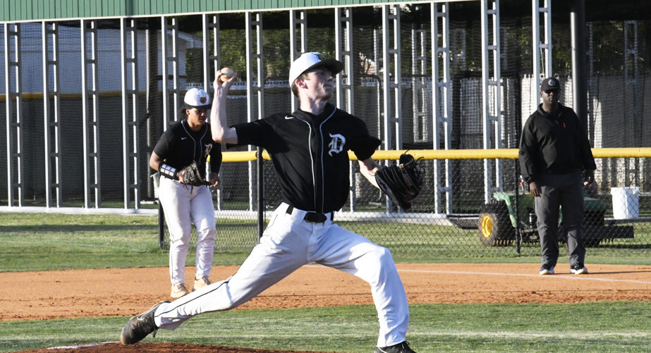 A baseball pitcher in a black and white uniform winds up to throw, with teammates and a coach visible in the background on a sunny day.