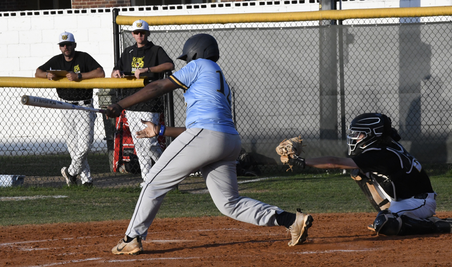 A baseball player in a blue jersey swings at a pitch while a catcher in black gear reaches to catch the ball.