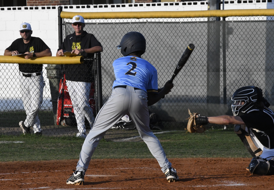 A baseball player in a blue jersey prepares to bat while a catcher squats behind him; two coaches watch from behind the fence.