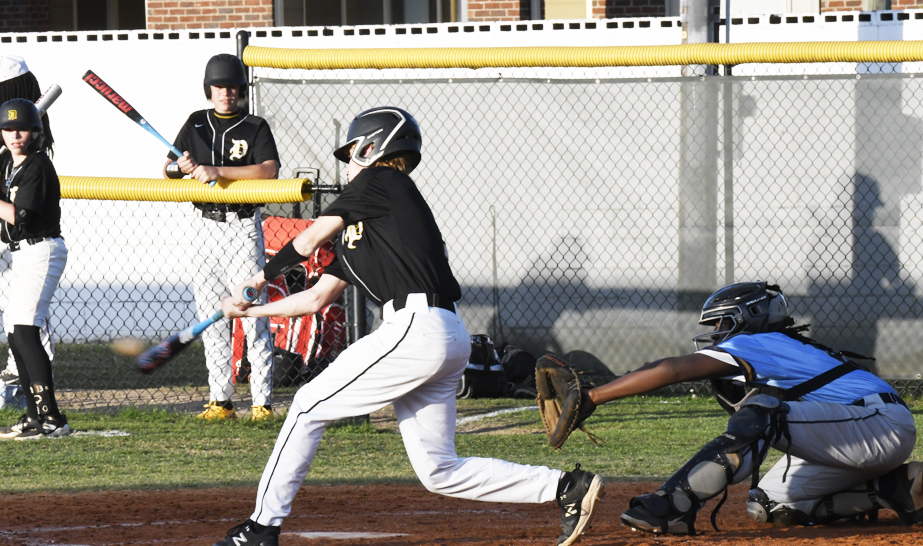 A youth baseball batter hits the ball as the catcher prepares to catch, with teammates watching from behind the fence.