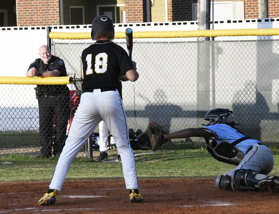 A youth baseball player at bat faces a catcher in blue during a game, with a person standing behind a chain-link fence in the background.