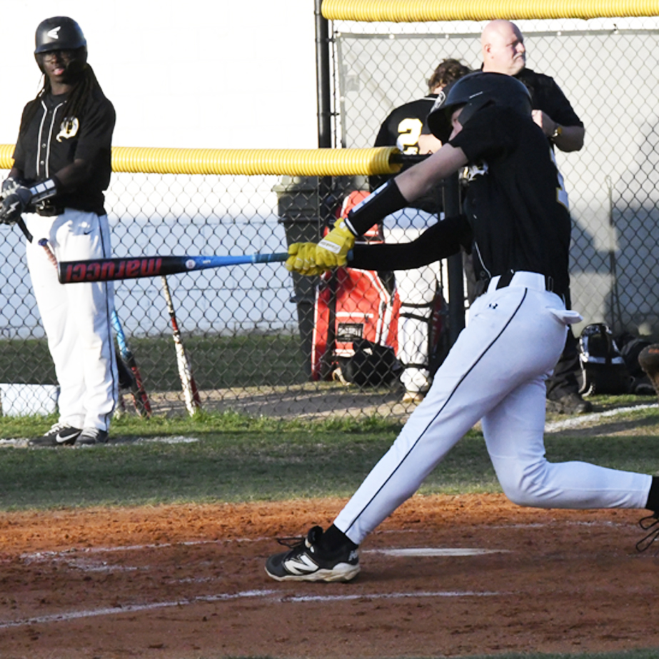 A baseball player swings a bat during a game, while another player and coach watch from behind a fence in the dugout area.