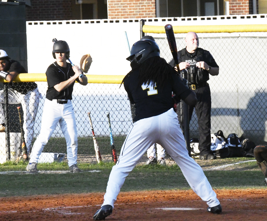 A baseball batter prepares to swing as the catcher and umpire watch, with a police officer standing in the background near the fence.