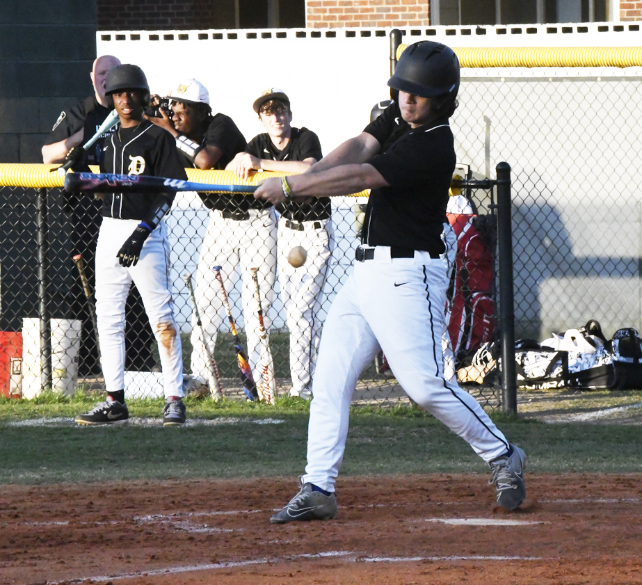 A baseball player in a black jersey swings at a pitch while teammates and coaches watch from behind a chain-link fence.