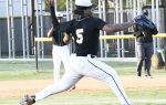 Baseball pitcher in black jersey with number 5 winding up to throw a pitch on a baseball field, with teammates and coach in the background.