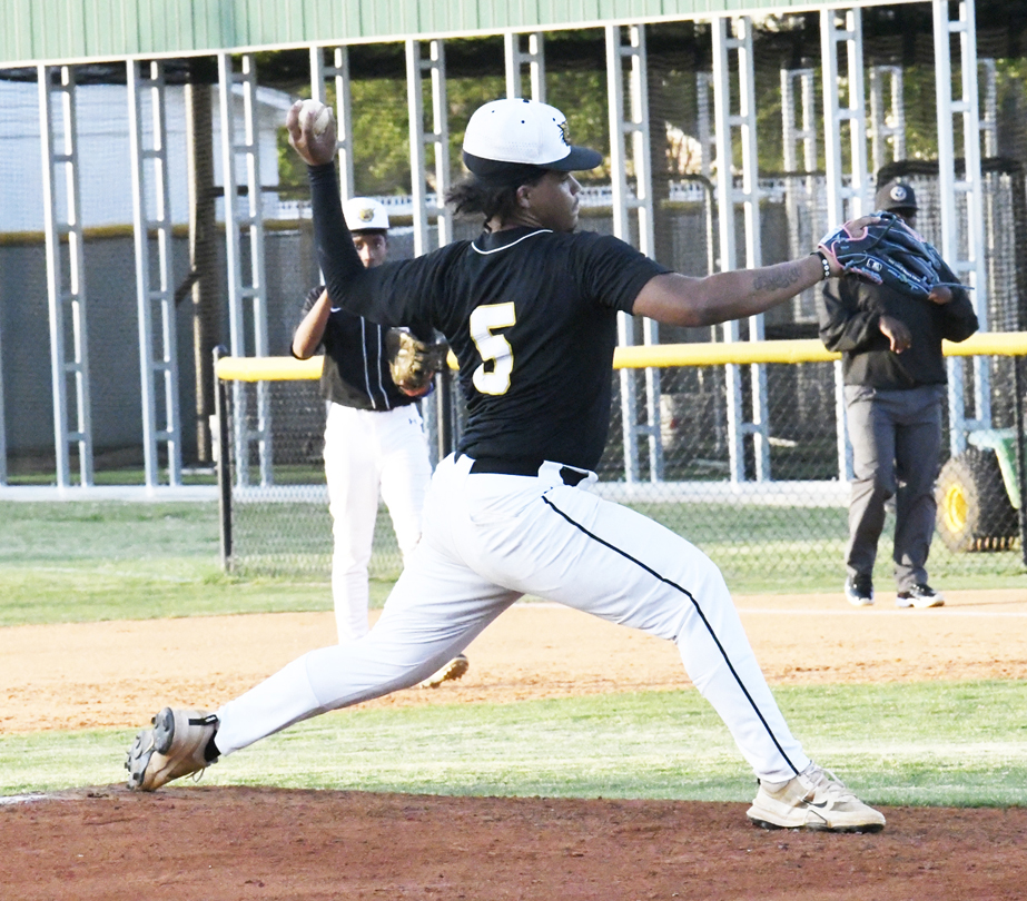 Baseball pitcher in black jersey with number 5 winding up to throw a pitch on a baseball field, with teammates and coach in the background.