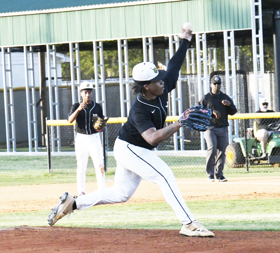 A baseball pitcher in black and white uniform throwing a pitch, with teammates and an umpire visible in the background on a sunny field.