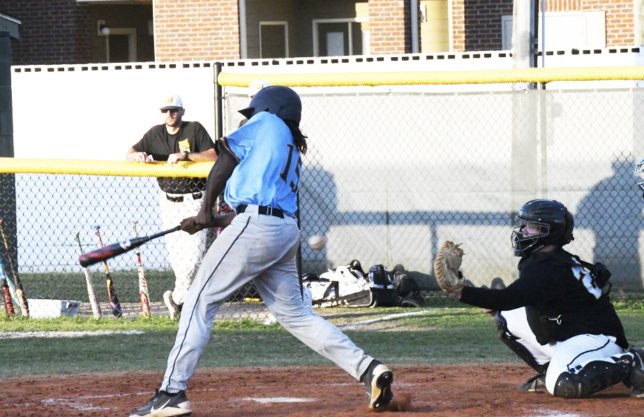 A baseball player swings at a pitch, with a catcher and umpire behind home plate, during a daytime game.