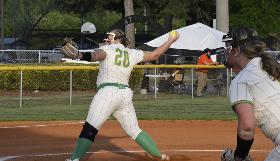 A softball pitcher in a white and green uniform, wearing number 20, throws a pitch while another player prepares to field on a dirt field.