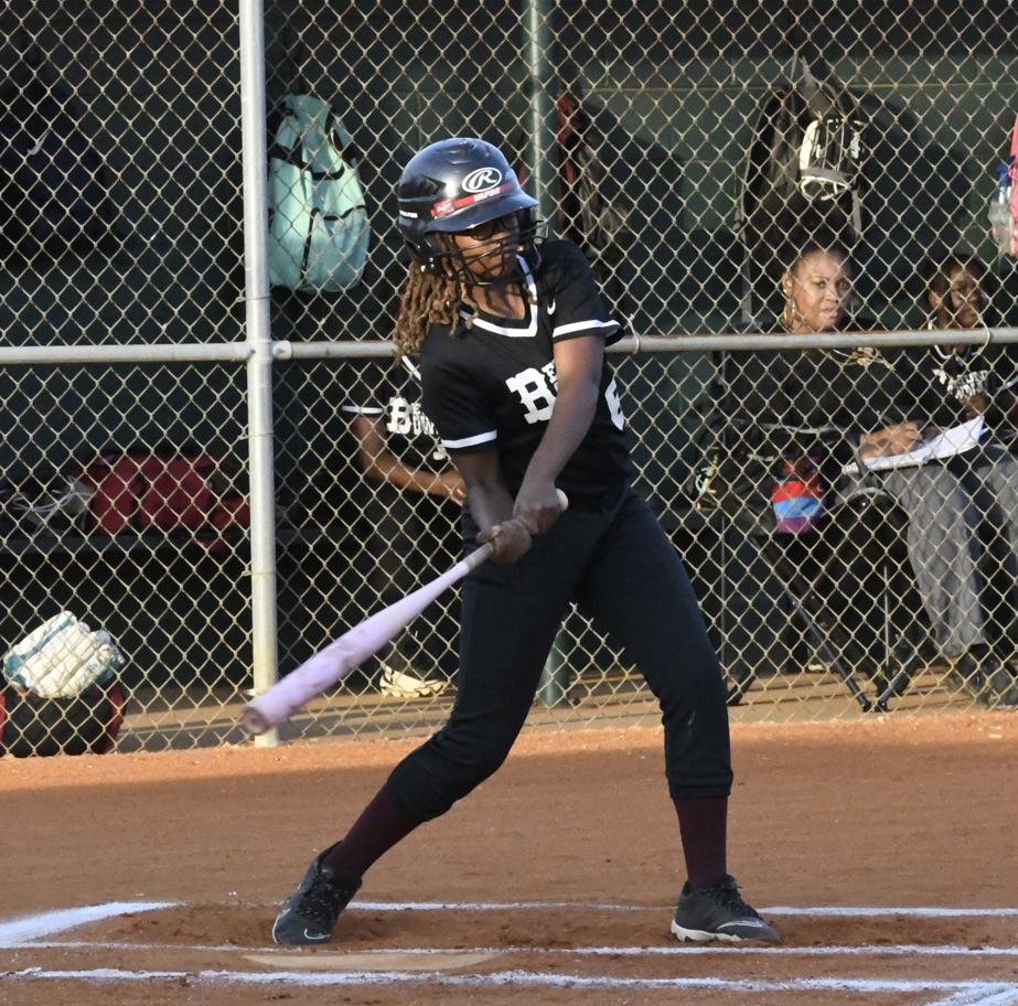 A softball player in black uniform swings a bat at home plate, with teammates and spectators behind a chain-link fence in the background.