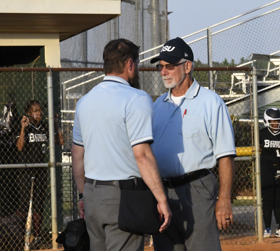 Two baseball umpires in light blue shirts and grey pants stand facing each other on the field, with players and a chain-link fence in the background.