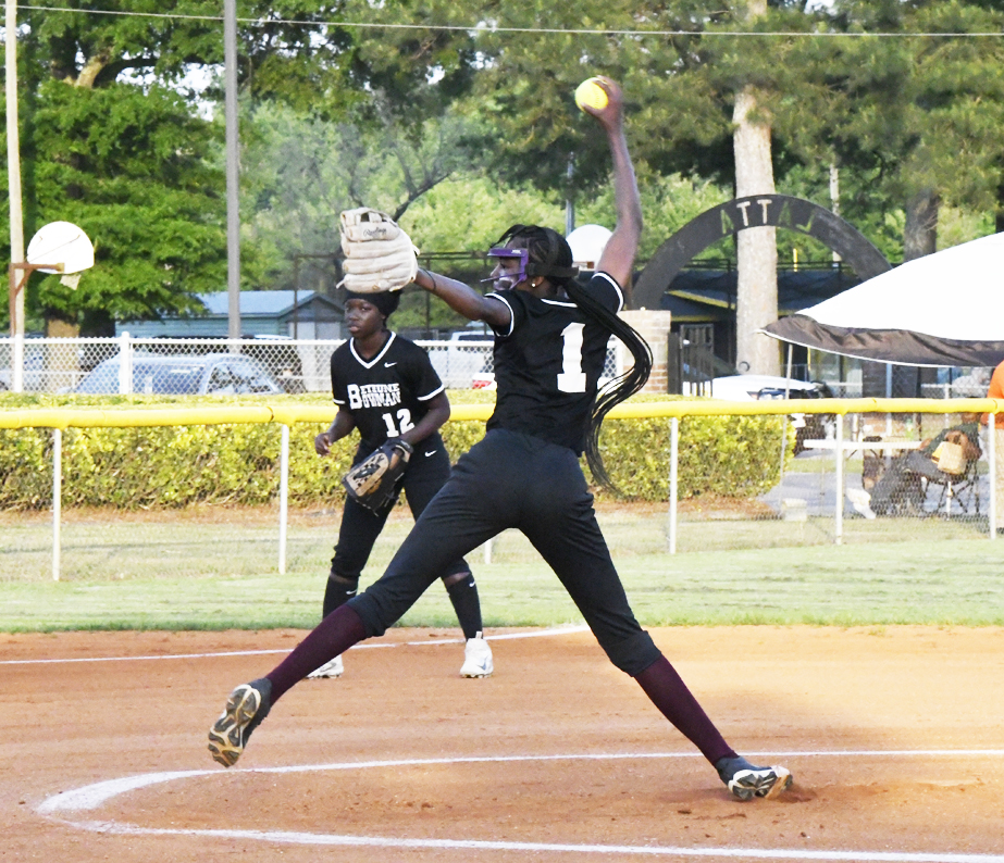 A softball pitcher in black uniform, mid-pitch, with a teammate in the background on a sunny outdoor field.