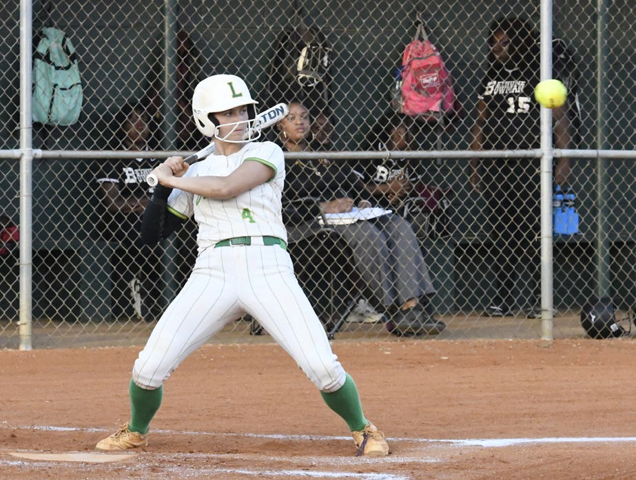 A softball player in white uniform prepares to bat as the ball approaches, with teammates and backpacks visible in the fenced dugout behind her.