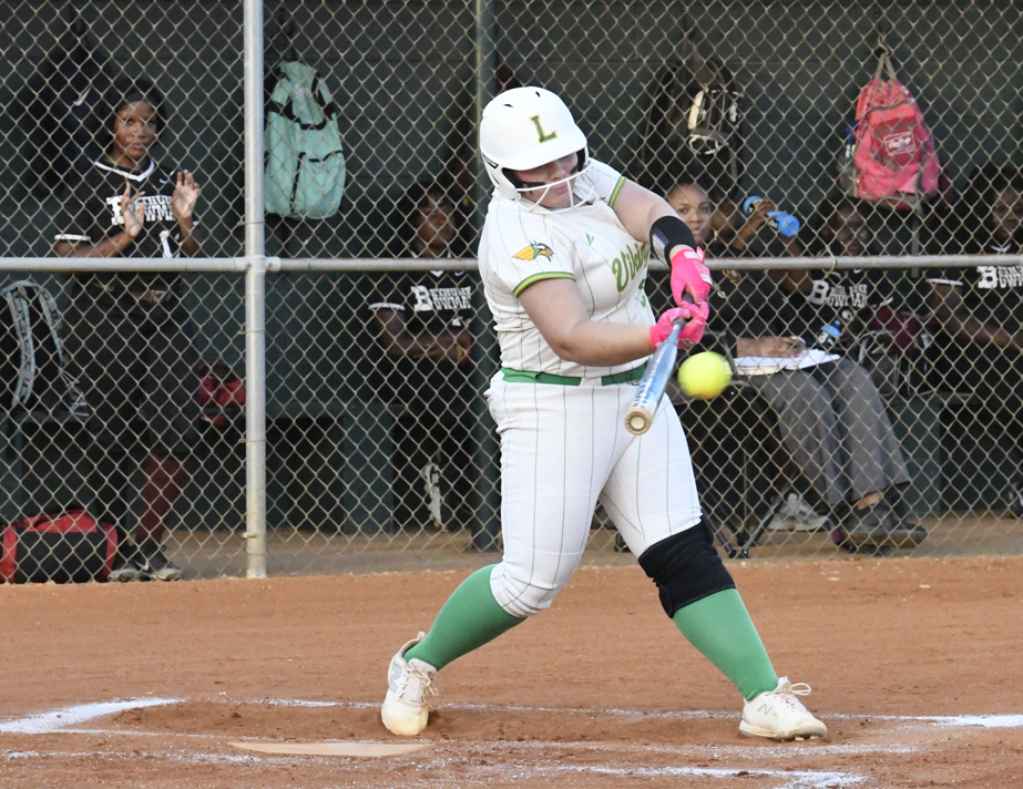 Softball player in white uniform hits the ball with a bat, while teammates and spectators watch from behind the fence in the dugout.