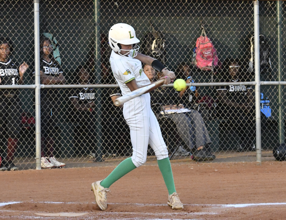 A softball player in white and green uniform hits a yellow ball during a game, with teammates and equipment bags visible behind a chain-link fence.