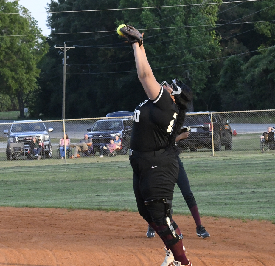 A softball catcher in black gear jumps and stretches upward to catch a ball during a game, with spectators and parked cars in the background.