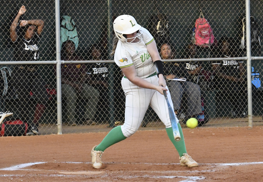 Softball player in white uniform swinging bat, hitting a pitch. Teammates sit and watch behind chain-link fence in dugout. Dirt infield and sports equipment visible.