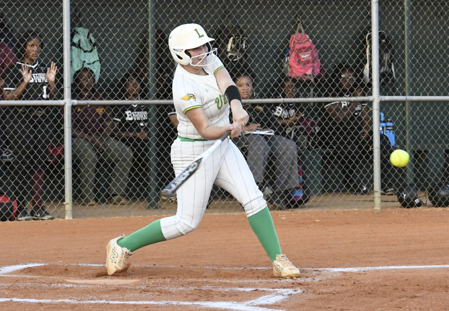 A softball player in a white and green uniform swings at a yellow ball, while teammates watch from the dugout behind a chain-link fence.