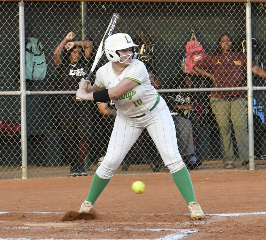 A softball player in white uniform prepares to swing at an incoming pitch, with teammates and spectators watching from behind a chain-link fence.